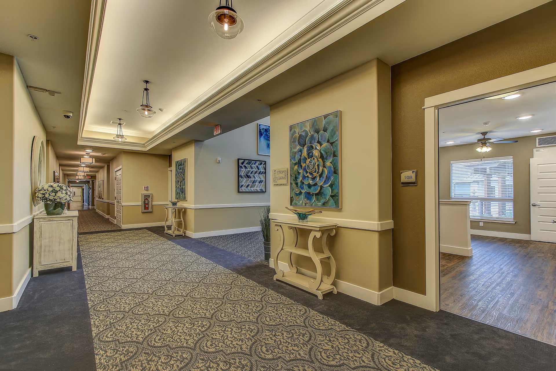 A well-lit hallway in a senior living facility with patterned carpet, beige walls, and decorative tables with artwork and plants. To the right, an open doorway leads to a room with wood flooring, a ceiling fan, and large windows with blinds.
