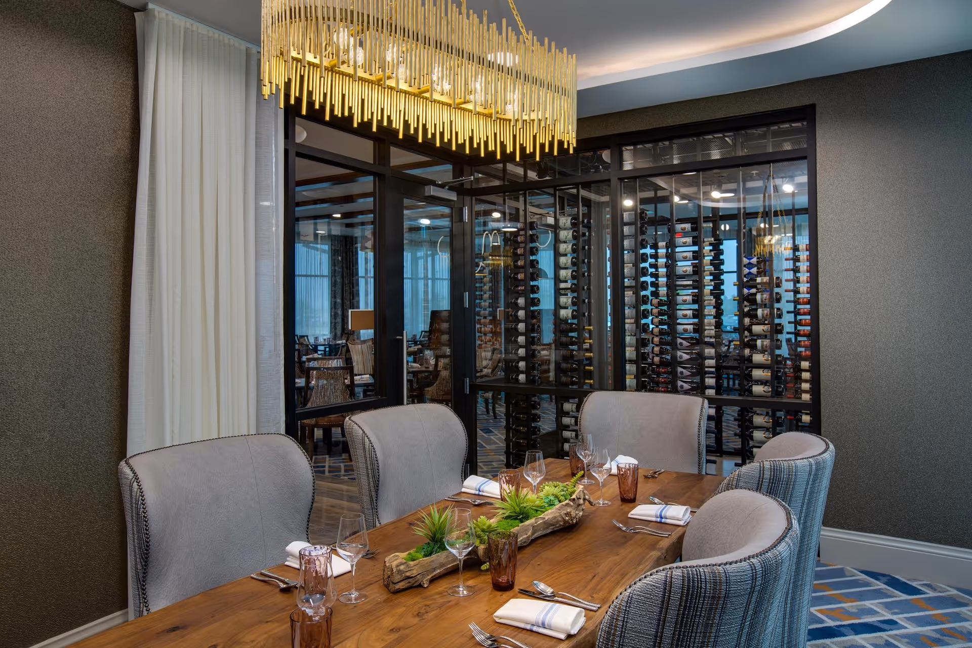 A modern dining room with a wooden table set for six people, featuring gray upholstered chairs and a centerpiece with green plants. Behind the table is a glass-enclosed wine storage area with multiple bottles displayed on racks. A large, contemporary gold chandelier hangs above the table, and the room has a patterned carpet and neutral-colored walls.