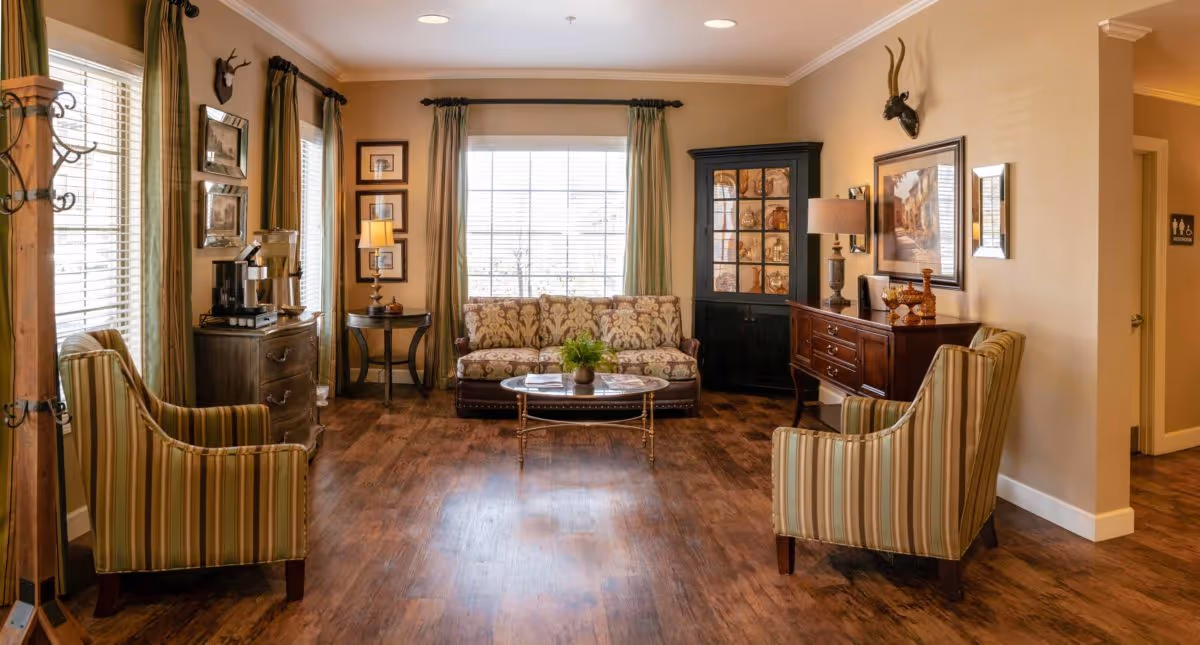 A cozy living room area with wooden flooring, a patterned sofa against a window with green curtains, two striped armchairs facing each other, a glass coffee table with a small plant, a wooden sideboard with a lamp and decorative items, framed artwork on the walls, and a black cabinet with glass doors. There is a coat rack on the left side and a hallway leading to a restroom on the right.