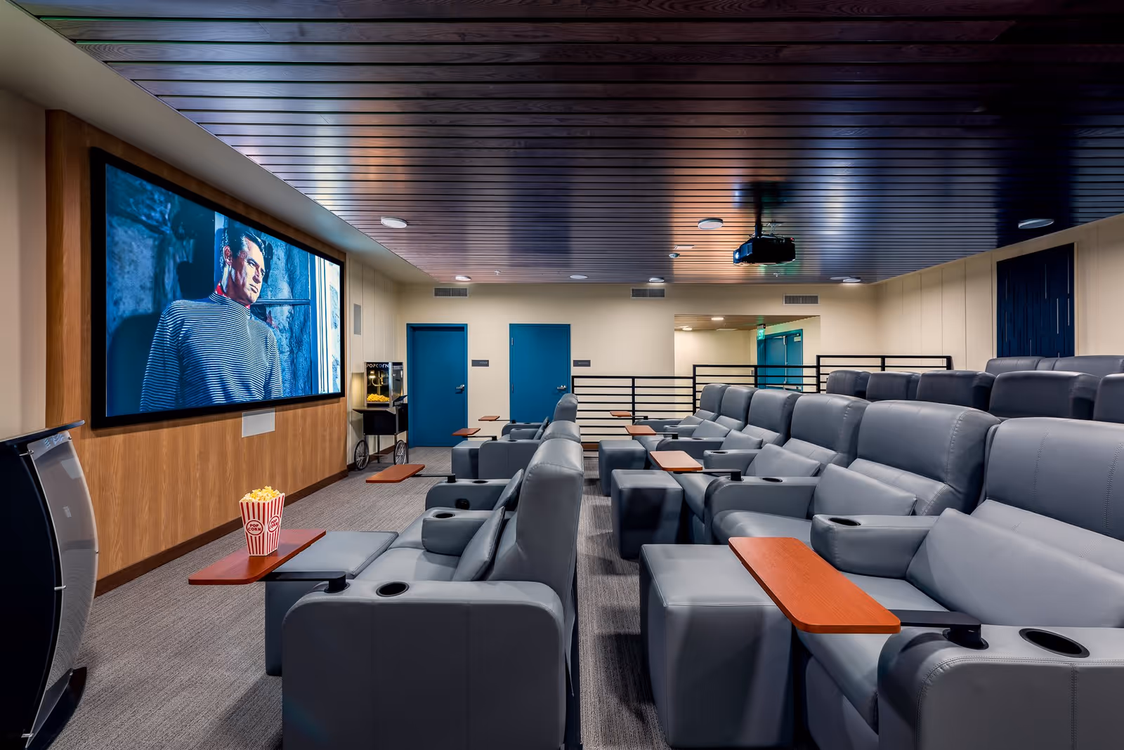 A modern theater room with multiple rows of gray leather reclining seats, each equipped with wooden trays and cup holders. A large screen on the wall displays a movie scene, and there is a popcorn machine near the back wall. The ceiling is wooden with recessed lighting and a projector mounted in the center.