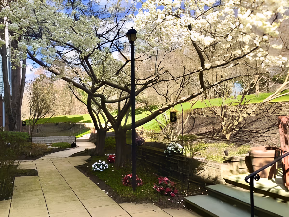 Outdoor pathway lined with blooming trees and flower beds, a lamppost in the center, steps with a railing on the right, and green grassy areas under a blue sky with some clouds.