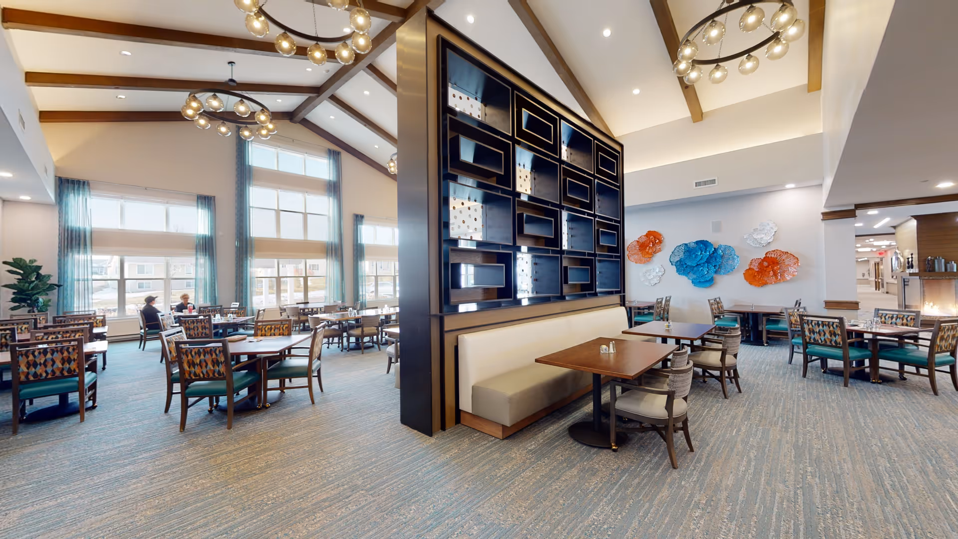 Bright, spacious dining room with multiple tables and chairs, vaulted ceilings, a central shelving partition, and colorful wall sculptures.