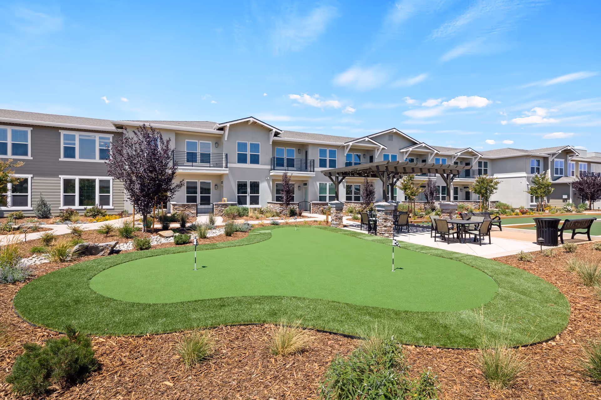 Outdoor view of Oakmont of Lodi senior living facility featuring a putting green with two golf holes, surrounded by landscaped garden beds with shrubs and trees. There is a pergola with seating and tables on a paved area adjacent to the green, and a two-story building with balconies and multiple windows in the background under a blue sky with scattered clouds.