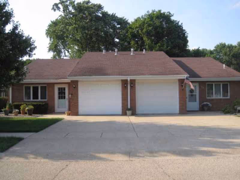 Front exterior view of a single-story brick building with two garage doors, two entry doors, and several windows. There are potted plants near the doors and an American flag hanging near the right door. Trees are visible in the background.