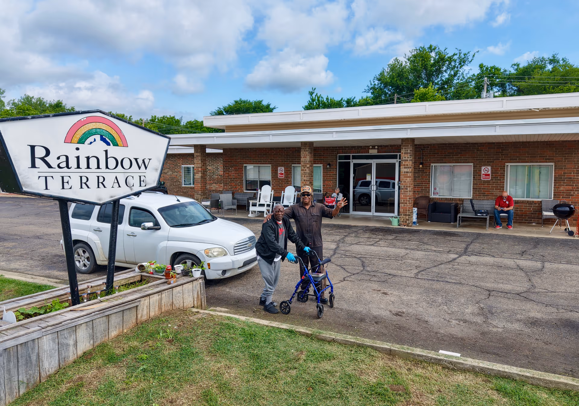 Exterior view of Rainbow Terrace Care Center showing a brick building with a covered entrance. Two elderly men are standing and smiling near a white vehicle, one using a walker. Another person is seated on a bench near the entrance. A large sign with a rainbow and the text 'Rainbow Terrace' is visible in the foreground.