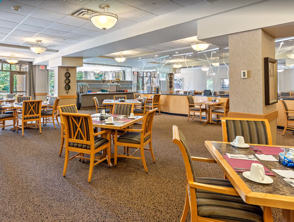 A spacious dining room with multiple wooden tables and chairs arranged neatly. Each table is set with placemats, cups, and utensils. The room has carpeted flooring, large windows allowing natural light, and ceiling lights. There is a serving area with a glass sneeze guard in the background.