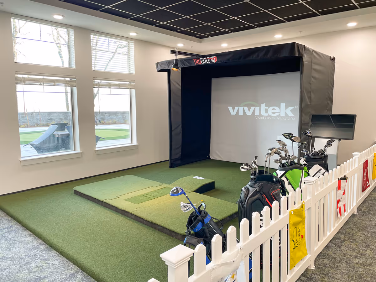 Indoor golf simulator area with a Vivitek projector screen, golf mats, and several golf bags with clubs behind a white picket fence. Large windows allow natural light into the room, and there is a monitor on a stand next to the simulator.