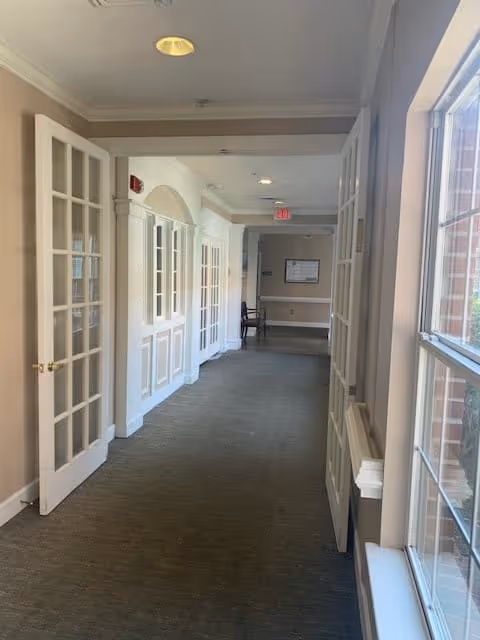 A carpeted hallway in an assisted living facility with beige walls and white trim. There are glass-paneled double doors on the left side and large windows on the right side letting in natural light. At the end of the hallway, there is a chair and a framed notice on the wall.