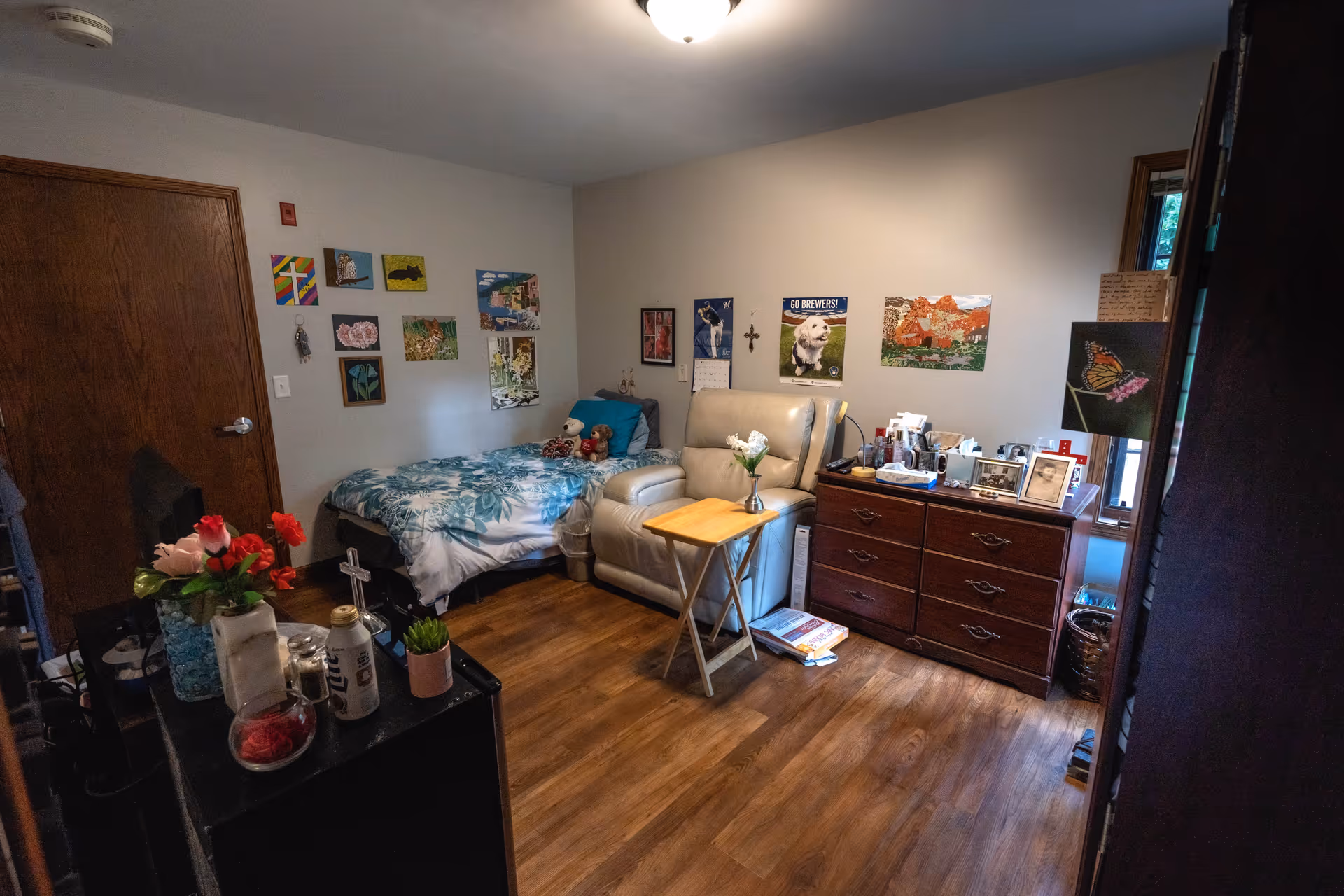 A cozy room featuring a bed with floral bedding, a comfortable chair, a small table, and various decorations on the walls, including paintings and photographs.