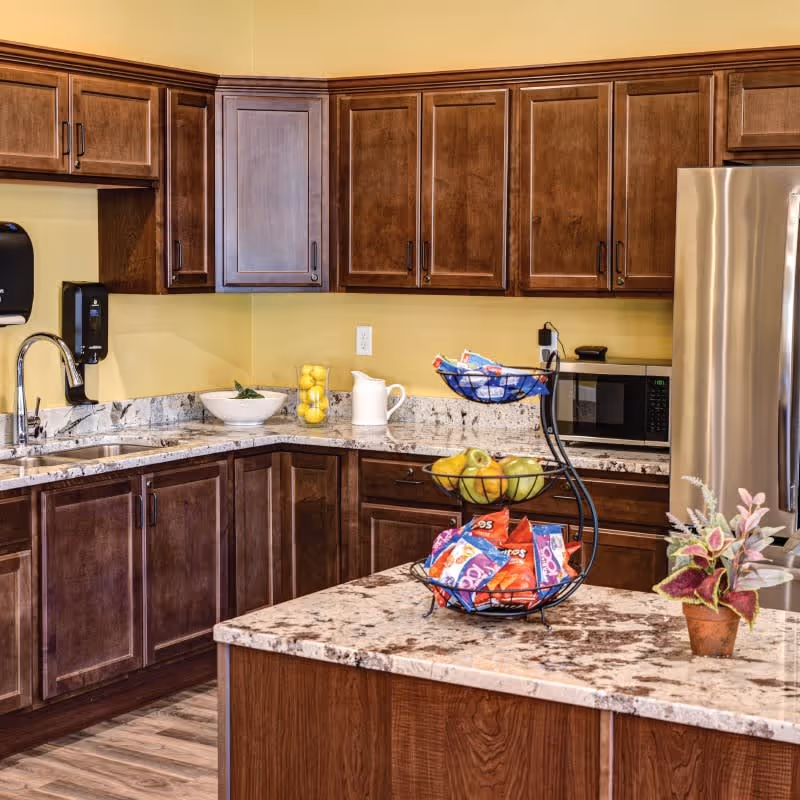 A kitchen area with dark wooden cabinets, a granite countertop island with a two-tiered fruit and snack basket, a stainless steel refrigerator, a microwave, a sink with a faucet, and a small potted plant on the island. The walls are painted yellow and the floor has wood-style flooring.