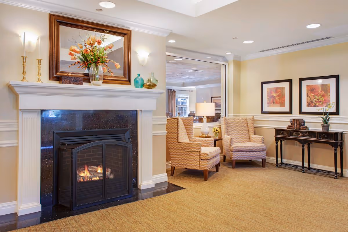 A cozy sitting area in a senior living facility featuring a lit fireplace with a white mantel decorated with a vase of flowers, candlesticks, and decorative glass bottles. Two patterned armchairs are positioned next to a side table with a lamp. On the wall behind the chairs are two framed floral artworks above a dark wooden console table with decorative items. The room has beige walls and carpeted flooring.
