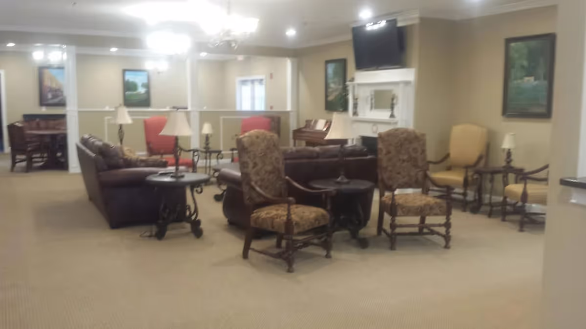 A spacious living room area in a senior living facility with multiple upholstered armchairs and sofas arranged around small round tables. The room features beige walls, carpeted floors, framed paintings, a piano, and a wall-mounted TV above a white fireplace mantel. Several table lamps provide additional lighting.