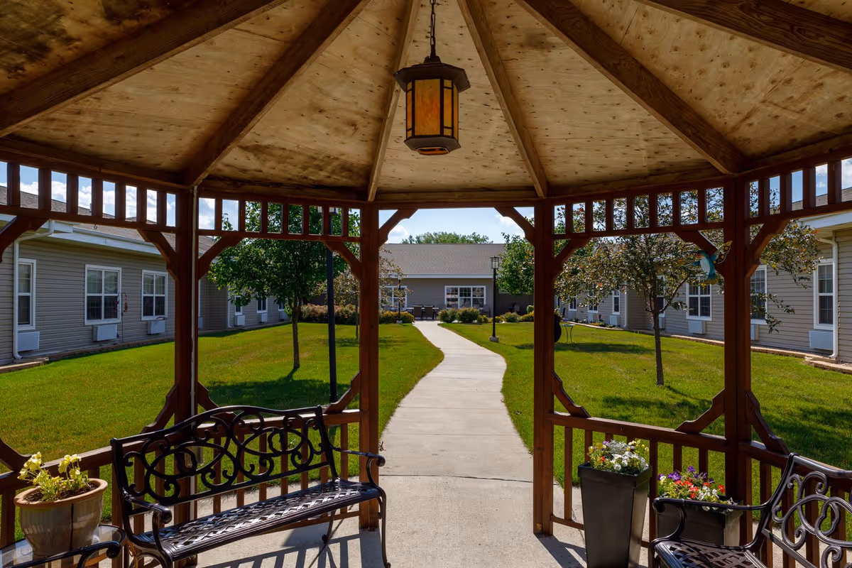 View from inside a wooden gazebo looking out onto a paved walkway leading through a grassy courtyard with small trees and shrubs, surrounded by single-story buildings with beige siding and white-trimmed windows. The gazebo has black metal benches and flower pots with colorful flowers.