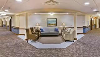 Carpeted senior living interior with a central seating area featuring a sofa, armchairs, lamps, and a framed painting, flanked by two corridors.