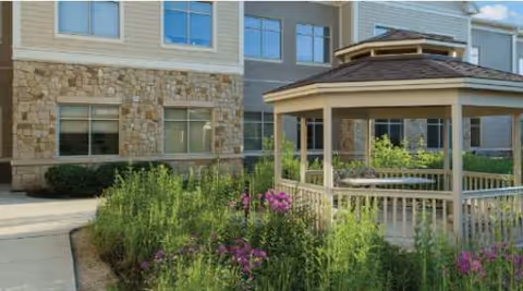 Outdoor garden area with a wooden gazebo surrounded by green plants and purple flowers, adjacent to a building with stone and siding exterior walls and multiple windows.