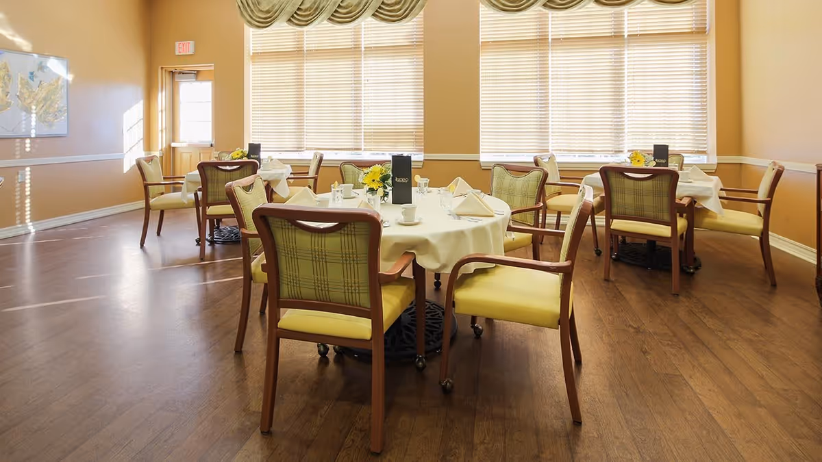 A bright dining room with several round tables covered with white tablecloths, each set with napkins, cups, and small flower arrangements. The room has wooden floors, large windows with blinds, and beige walls with a framed picture and an exit door in the background.