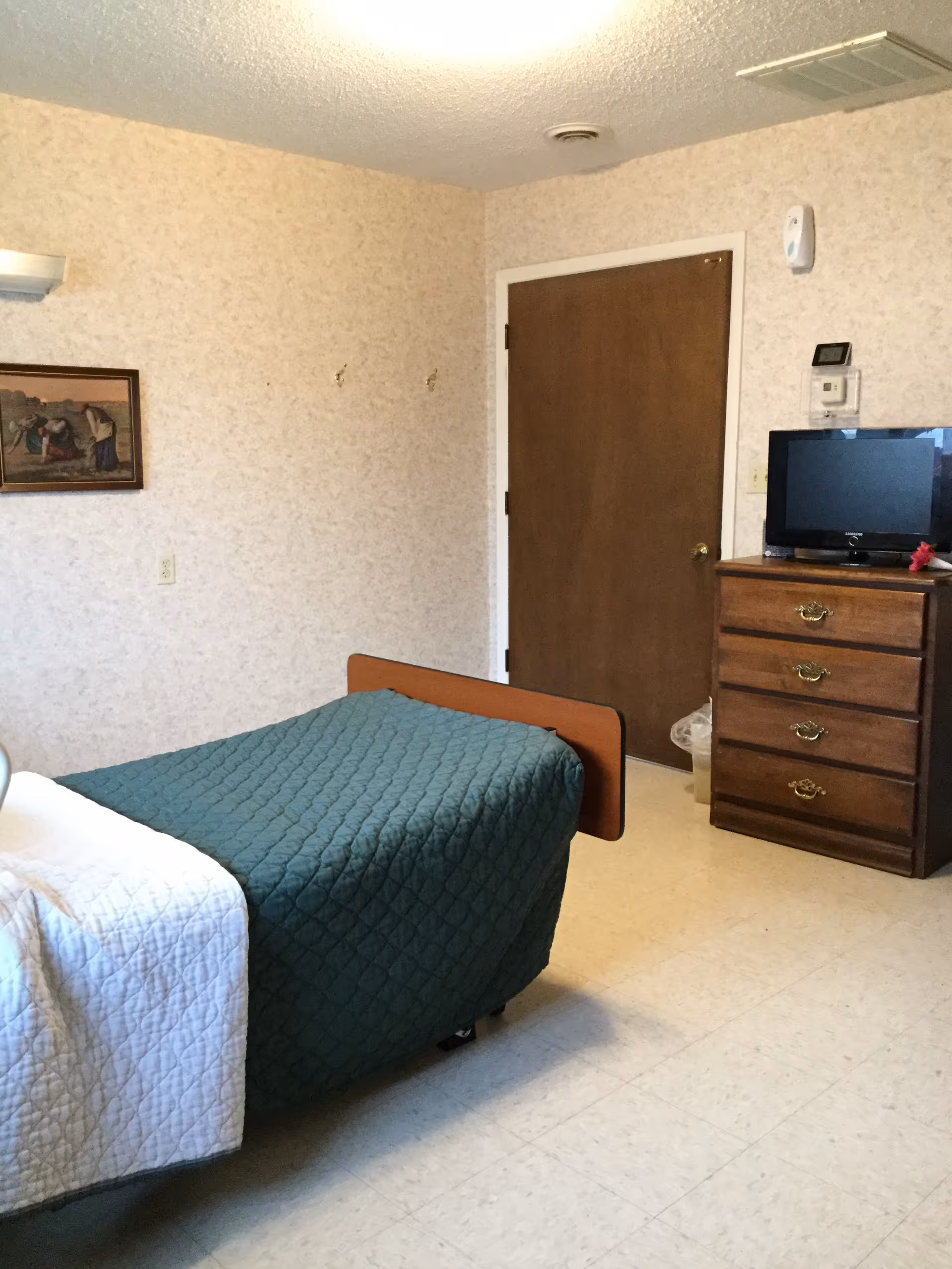 A simple bedroom with a single bed covered with a green quilt and a white blanket at the foot. There is a wooden dresser with a small television on top, a closed wooden door, and a framed picture on the wall. The walls have a light patterned wallpaper and the floor is tiled.