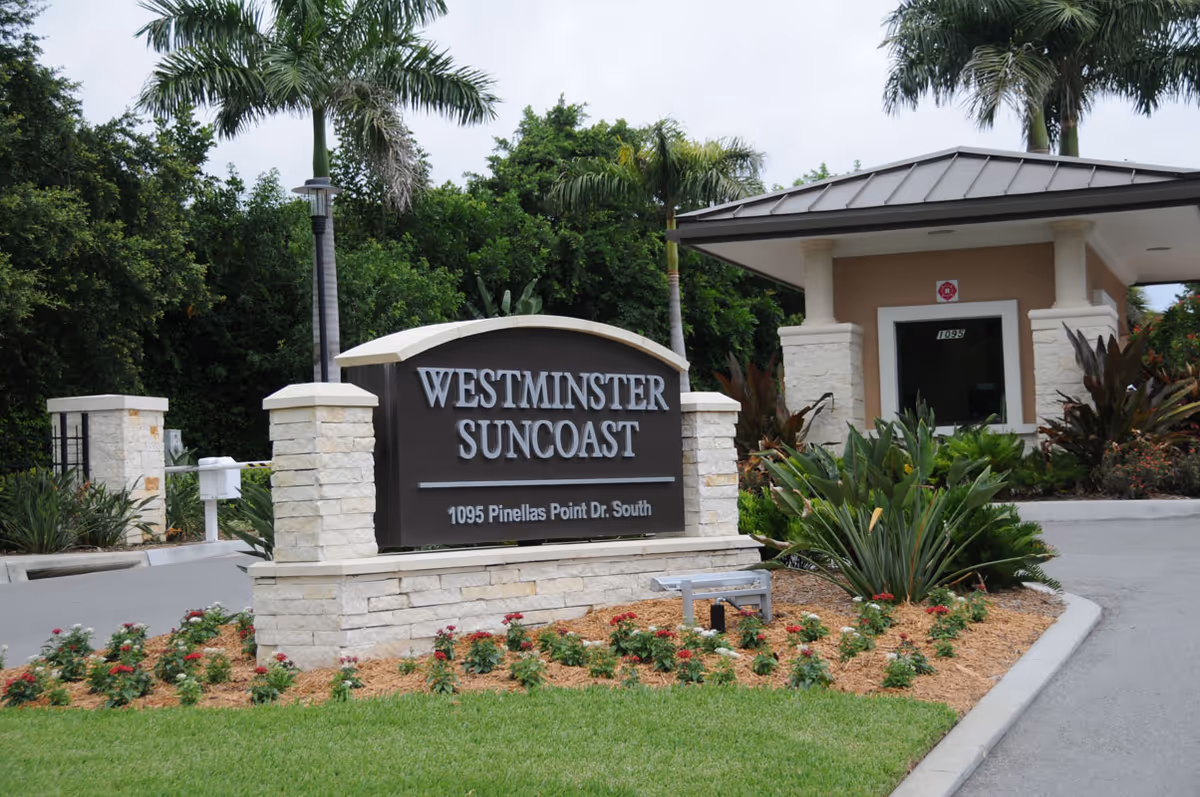Entrance sign for Westminster Suncoast facility with address 1095 Pinellas Point Dr. South, surrounded by landscaping including palm trees, bushes, and flowers, with a guardhouse visible in the background.