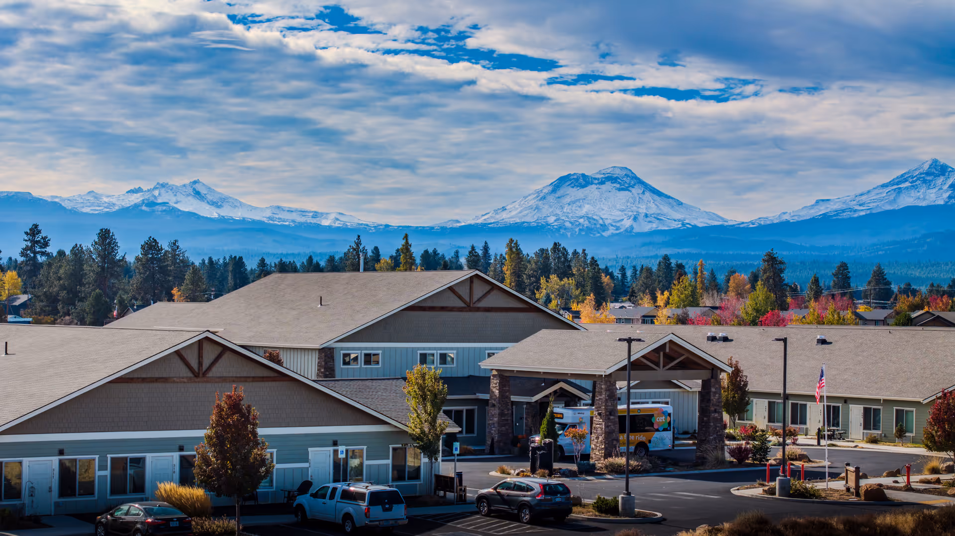 Exterior front view of The Lodge Retirement Community showing the entrance, parking lot, and snow-capped mountains beyond.
