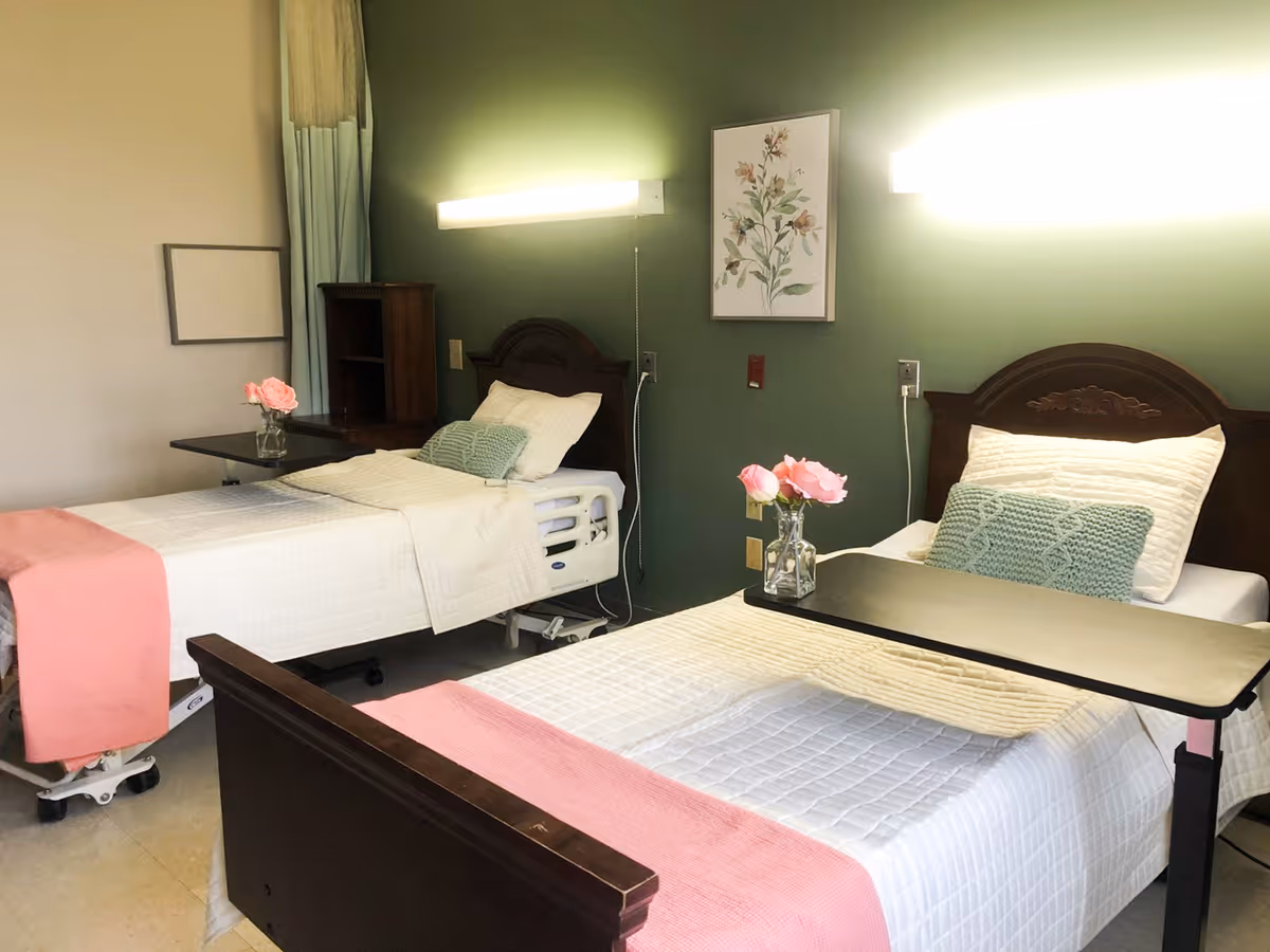 A room with two hospital-style beds, each with white bedding, a green pillow, and a pink throw blanket. Each bed has a small black overbed table with a vase holding pink flowers. The walls are painted green and beige, with a floral picture hanging on the green wall. There are wall-mounted lights above each bed and a wooden cabinet between the beds.