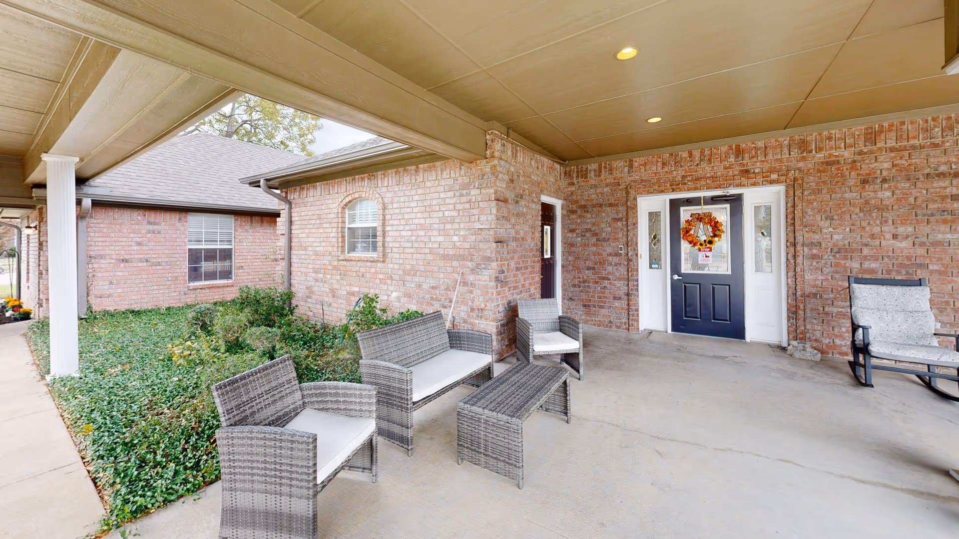 Covered entrance patio with wicker seating and a wreath-decorated front door on a brick senior living building.