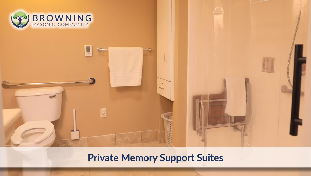 A bathroom with a toilet, a towel hanging on a wall-mounted rack, a grab bar next to the toilet, a shower area with a clear curtain, and a fold-down shower seat with a towel on it. The walls are beige and the floor is tiled.