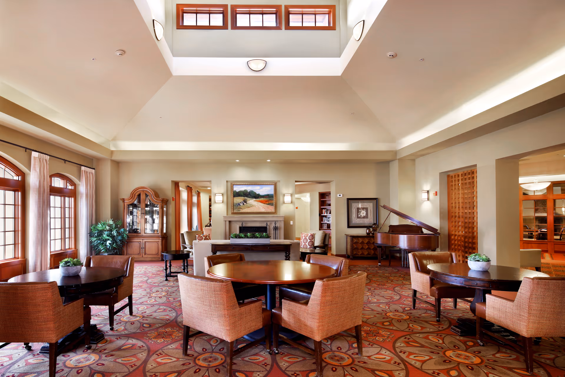 A spacious and well-lit living room area with high ceilings and multiple windows near the ceiling. The room features several round tables with cushioned chairs, a grand piano, a fireplace with a landscape painting above it, and a wooden cabinet with glass doors. The carpet has a colorful, intricate pattern, and there are plants placed on the tables and near the windows.