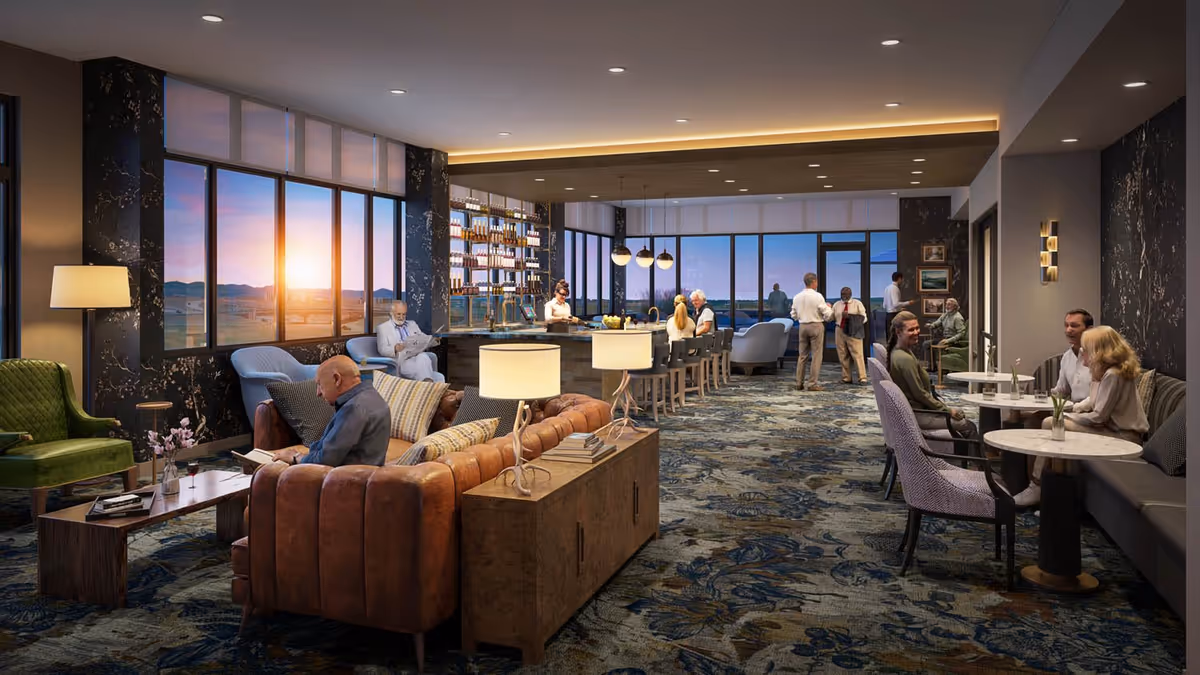A spacious, modern lounge area with large windows showing a sunset view. The room features comfortable seating including a leather sofa, armchairs, and small tables. There is a bar area with stools and shelves stocked with bottles. Several people are seated or standing, engaging in conversation or reading. The carpet has a floral pattern, and the lighting is warm and inviting.