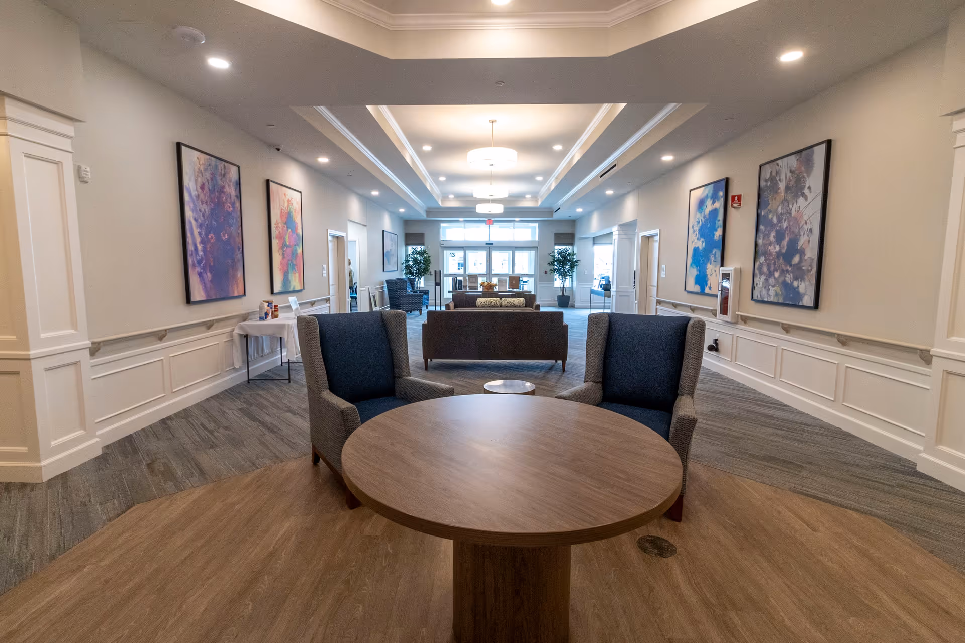 A spacious and well-lit common area in a senior living facility featuring a round wooden table with two upholstered armchairs in the foreground. The room has a high ceiling with recessed lighting and modern hanging light fixtures. The walls are adorned with colorful abstract paintings, and there are additional seating areas with sofas and chairs further back near large windows and glass doors that let in natural light. Potted plants are placed near the entrance, adding a touch of greenery.
