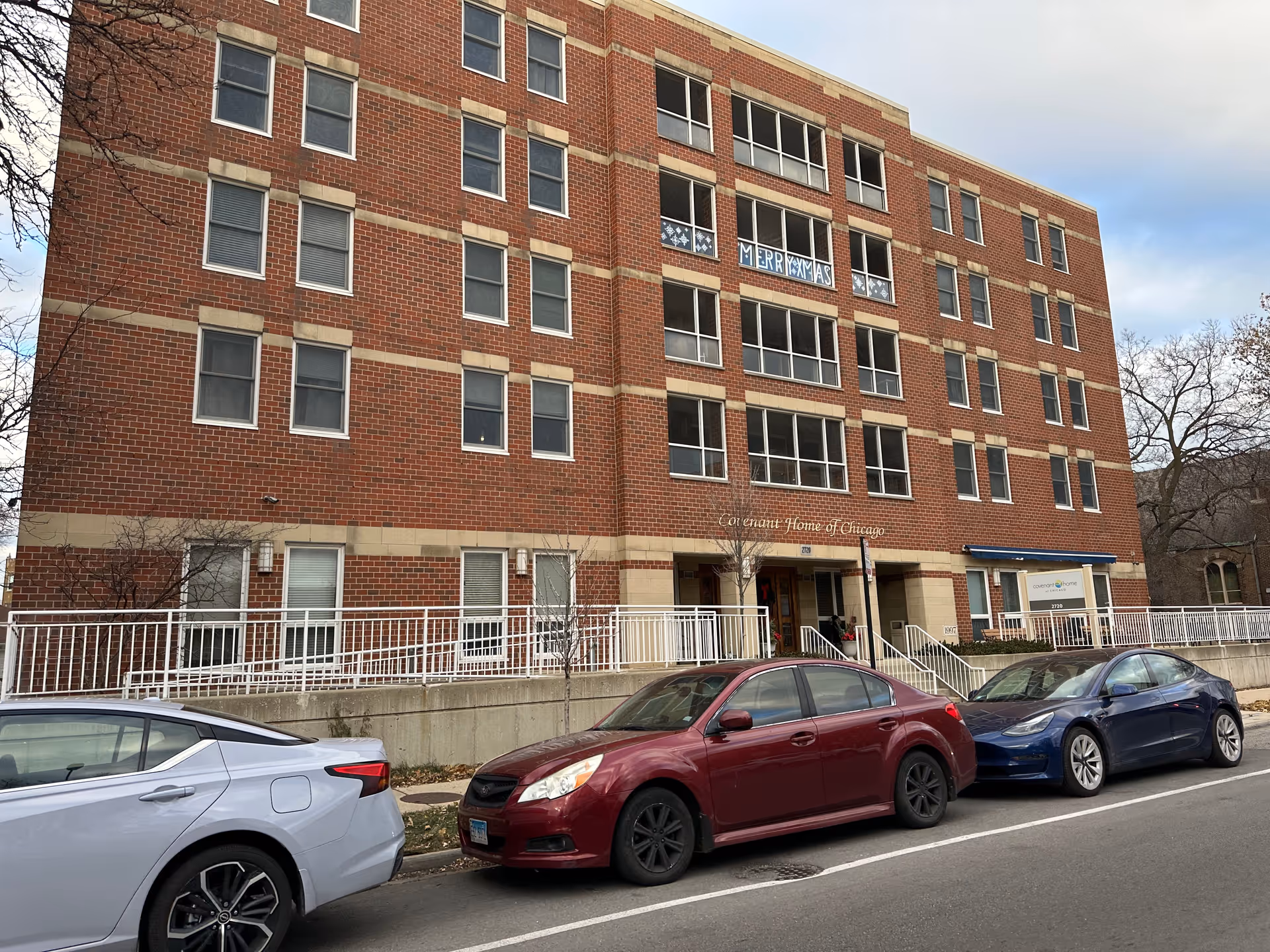 Exterior view of a four-story brick building with multiple windows and a sign that reads 'Covenant Home of Chicago' above the entrance. Three cars are parked along the street in front of the building. There is a festive 'Merry Xmas' decoration visible in one of the upper windows.