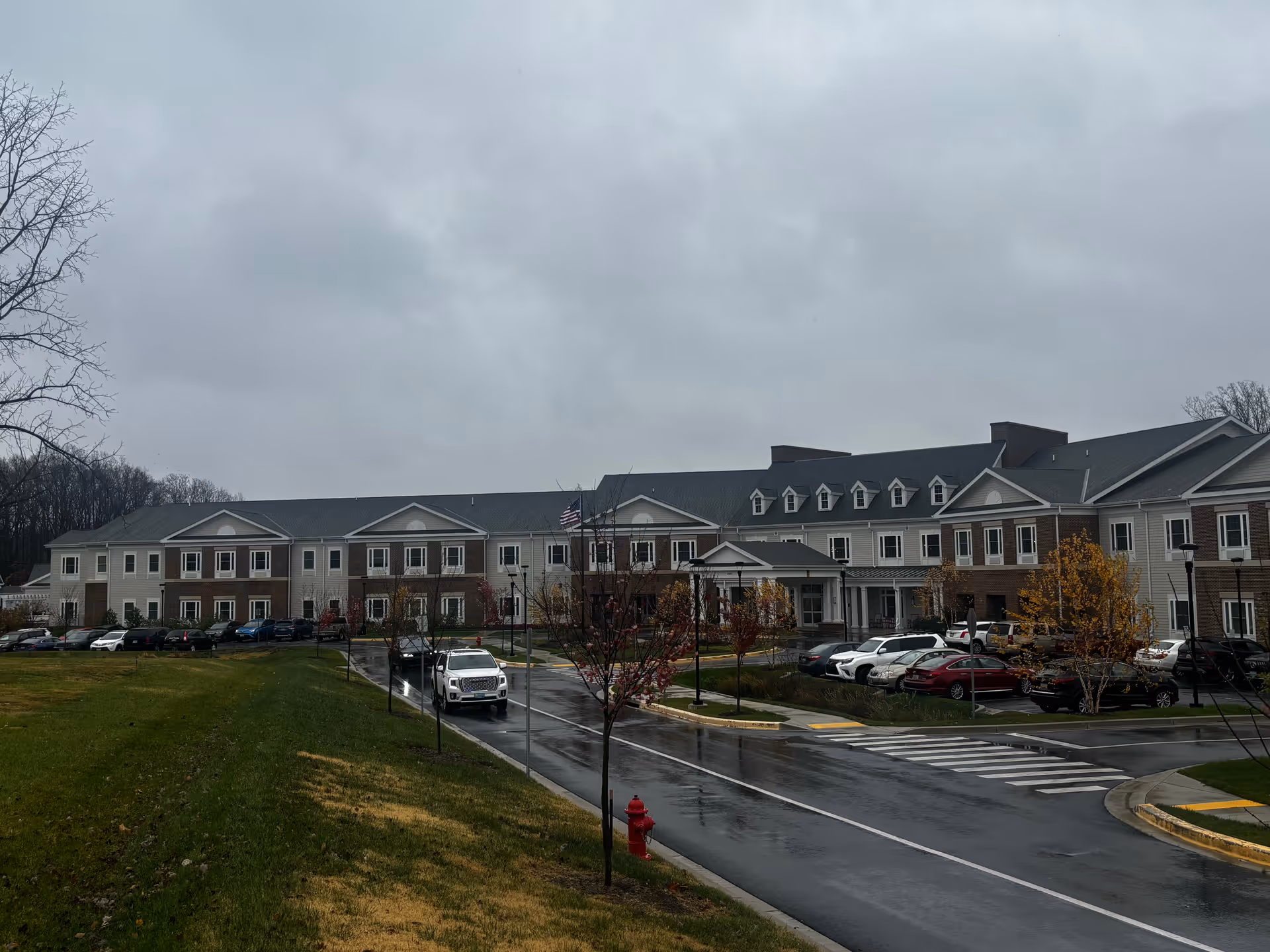 Exterior view of a large senior living facility building on a cloudy day with a wet road and parked cars in front. The building has multiple windows, a covered entrance, and an American flag flying near the center.