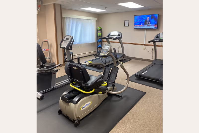 Indoor fitness room with exercise equipment including a recumbent NuStep machine, treadmills, and a rack of colorful dumbbells. A wall-mounted TV is showing a news channel. The room has beige walls, a window with vertical blinds, and carpeted flooring with black mats under the equipment.
