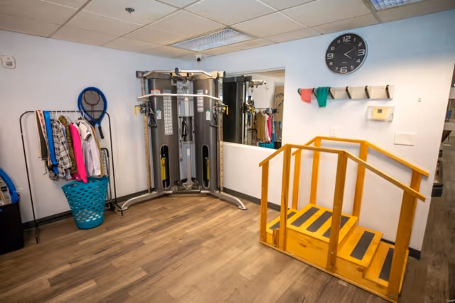 A rehabilitation or exercise room with a multi-functional gym machine, a small wooden staircase with handrails, a wall clock, a mirror, and a rack holding clothes and exercise bands.