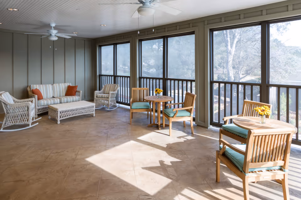 A bright, enclosed patio area with large screened windows allowing natural light to fill the space. The room features a wicker sofa with striped cushions and orange throw pillows, two wicker rocking chairs, a wicker coffee table, and two small wooden tables each with two wooden chairs with green cushions. Small vases with yellow flowers are placed on the tables. Ceiling fans are mounted on the white paneled ceiling.