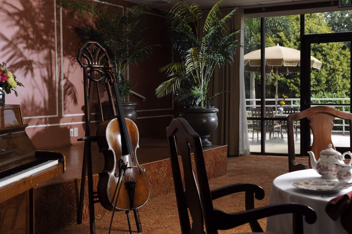 Interior room with a piano and a cello on a stand next to it, a table set with a teapot and cups, wooden chairs, large potted plants, and a glass door leading to an outdoor patio with tables, chairs, and umbrellas.