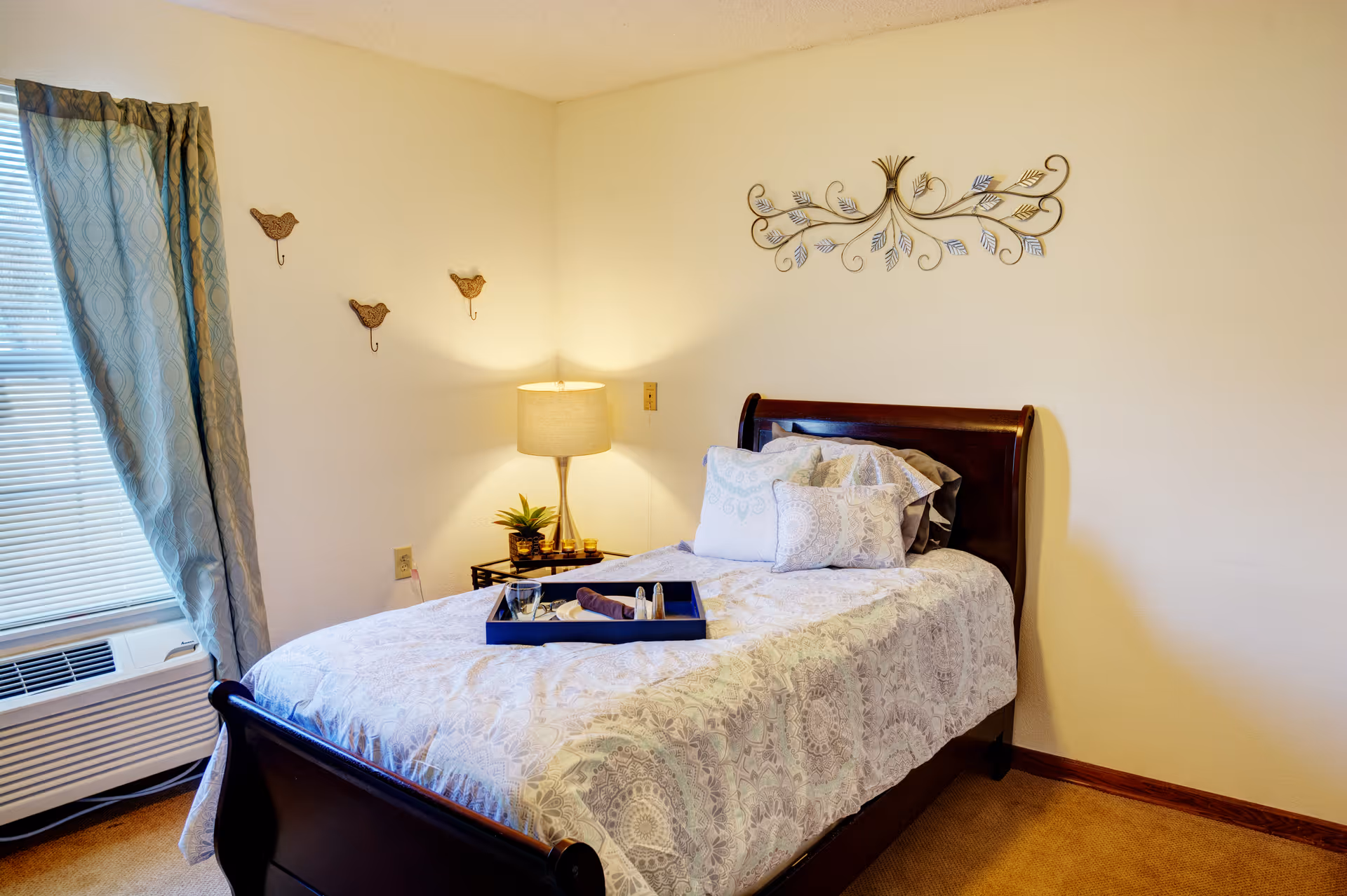 Well-lit bedroom with a single wooden bed dressed in patterned bedding, a nightstand with a lamp, window with curtains, and wall decorations.