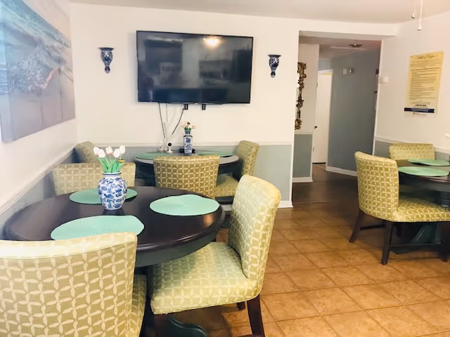 Communal dining area with round dark tables, green patterned chairs, placemats, a wall-mounted TV, and decorative vases.