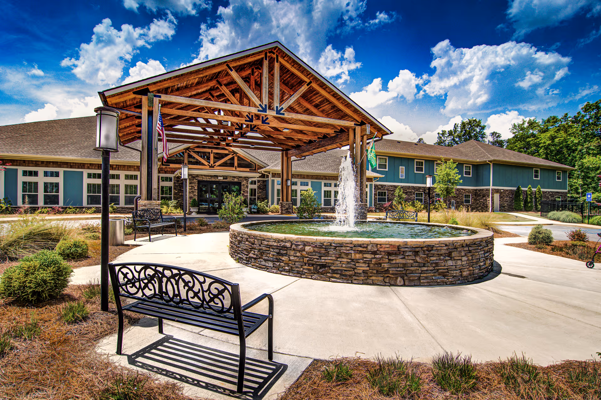 Outdoor view of Oaks at Grove Park facility entrance featuring a covered wooden porte-cochere, a circular stone fountain with water spraying upwards, black metal benches, landscaped greenery, and a partly cloudy blue sky.