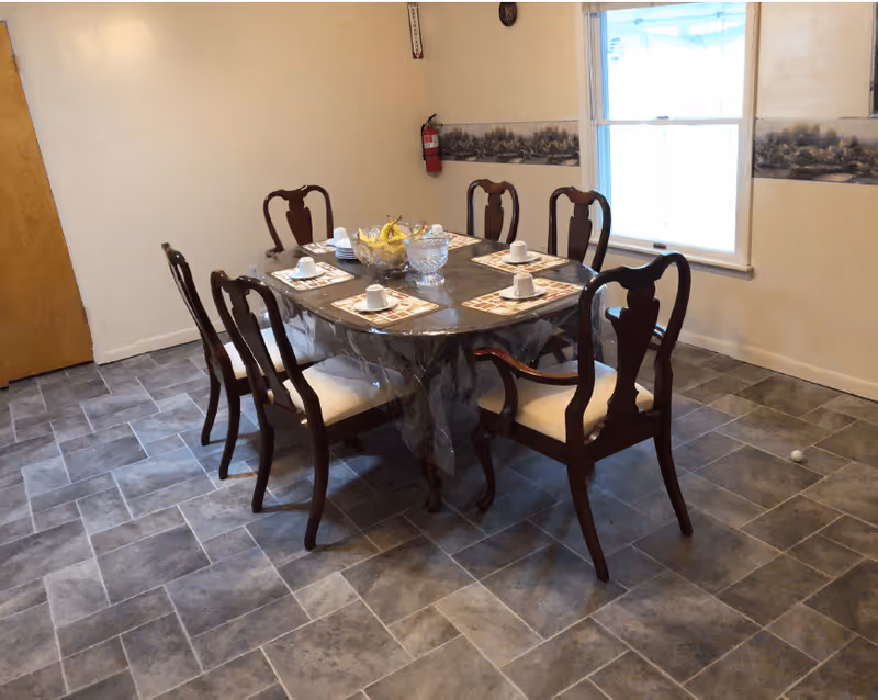 A dining room with a rectangular wooden table covered with a clear plastic tablecloth. Six wooden chairs with white cushions surround the table. The table is set with placemats, cups, and a centerpiece bowl with bananas. There is a window on the right wall and a fire extinguisher mounted on the back wall.