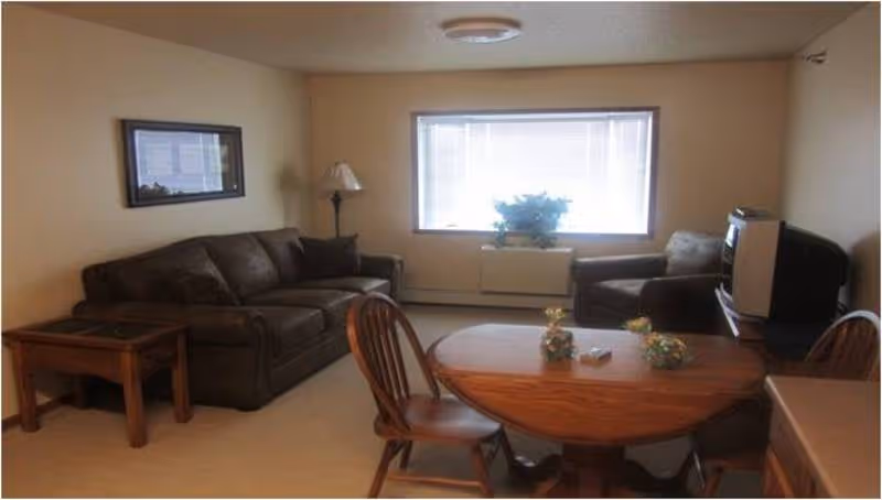 A living room with a brown sofa, an armchair, a wooden coffee table, and a wooden dining table with chairs. There is a floor lamp next to the sofa, a window with blinds, and a television on a stand.