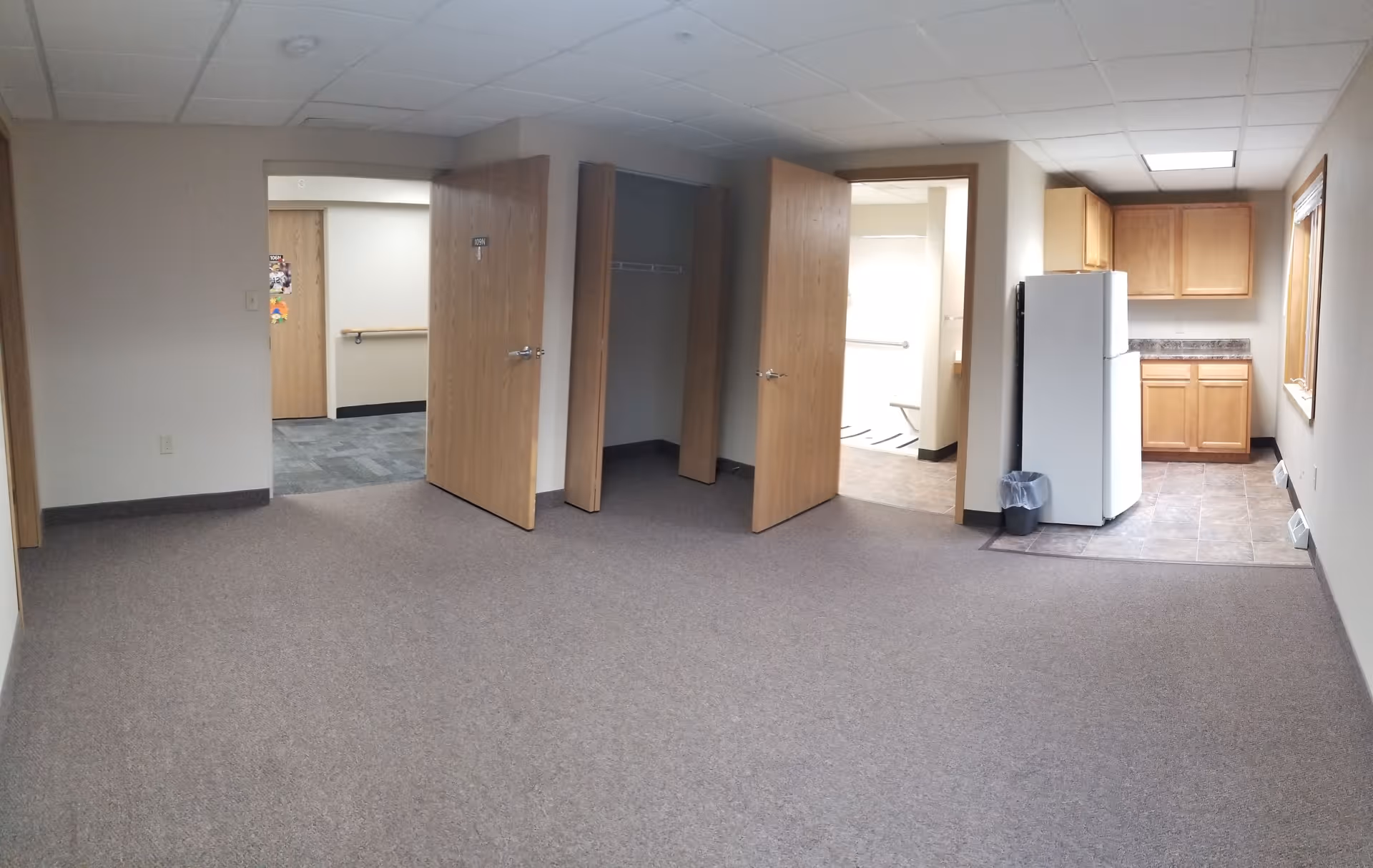 Interior view of a room with beige carpet and white walls. There are three wooden doors, one leading to a hallway with carpeted floor, one open closet door, and one door leading to a small kitchen area with a refrigerator, wooden cabinets, and tiled floor. The ceiling has white tiles with fluorescent lighting.