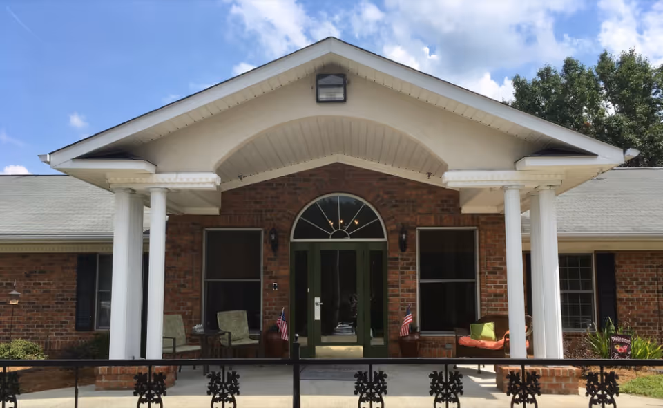 Front exterior view of a brick building with a green double door entrance, two white columns supporting a small roof over the entrance, two chairs on the porch, small American flags on either side of the door, and a welcome sign on the right side near some plants. The sky is partly cloudy.
