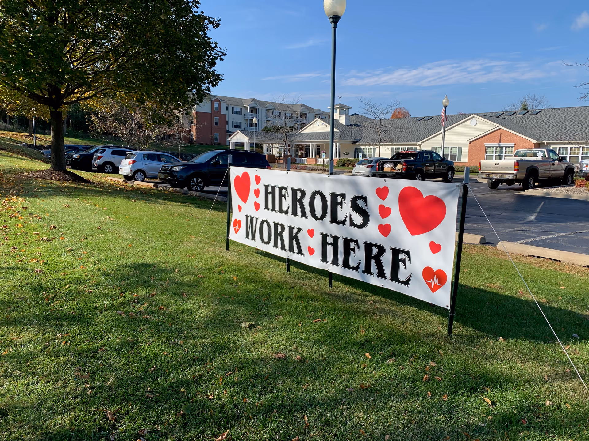 A lawn banner reading "HEROES WORK HERE" with red hearts on the grass in front of a senior living building and parked cars.