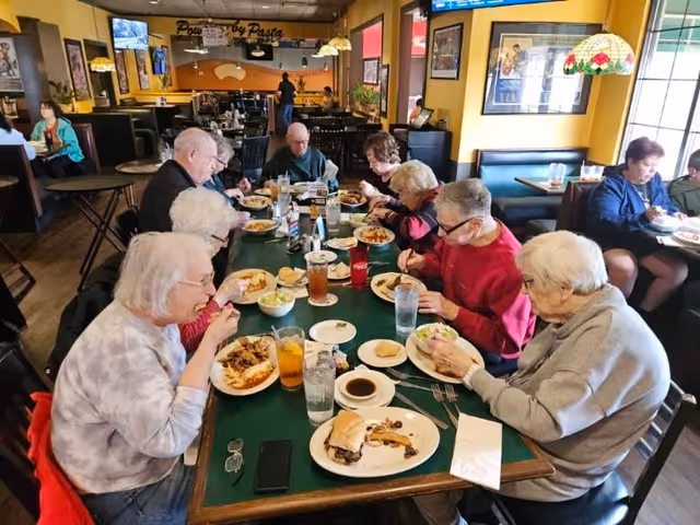 A group of elderly people sitting around a long dining table in a restaurant, eating various meals including sandwiches, salads, and pasta. The restaurant has yellow walls, framed pictures, and stained glass hanging lamps. Other diners are visible in the background.