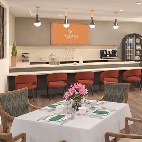 A dining area in Valage Senior Living At Carson Valley featuring a table set for four with white tablecloth, green napkins, cups, plates, and silverware. In the background, there is a counter with five red chairs, pendant lights hanging from the ceiling, a coffee machine, and a cabinet with shelves holding various items. A framed sign with the Valage Senior Living logo is mounted on the wall above the counter.