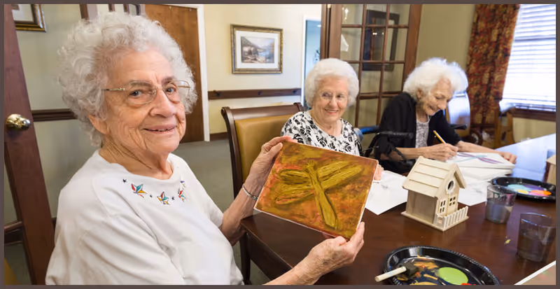 Three older women seated at a table in a communal room doing arts and crafts, with one woman holding up a painted canvas.