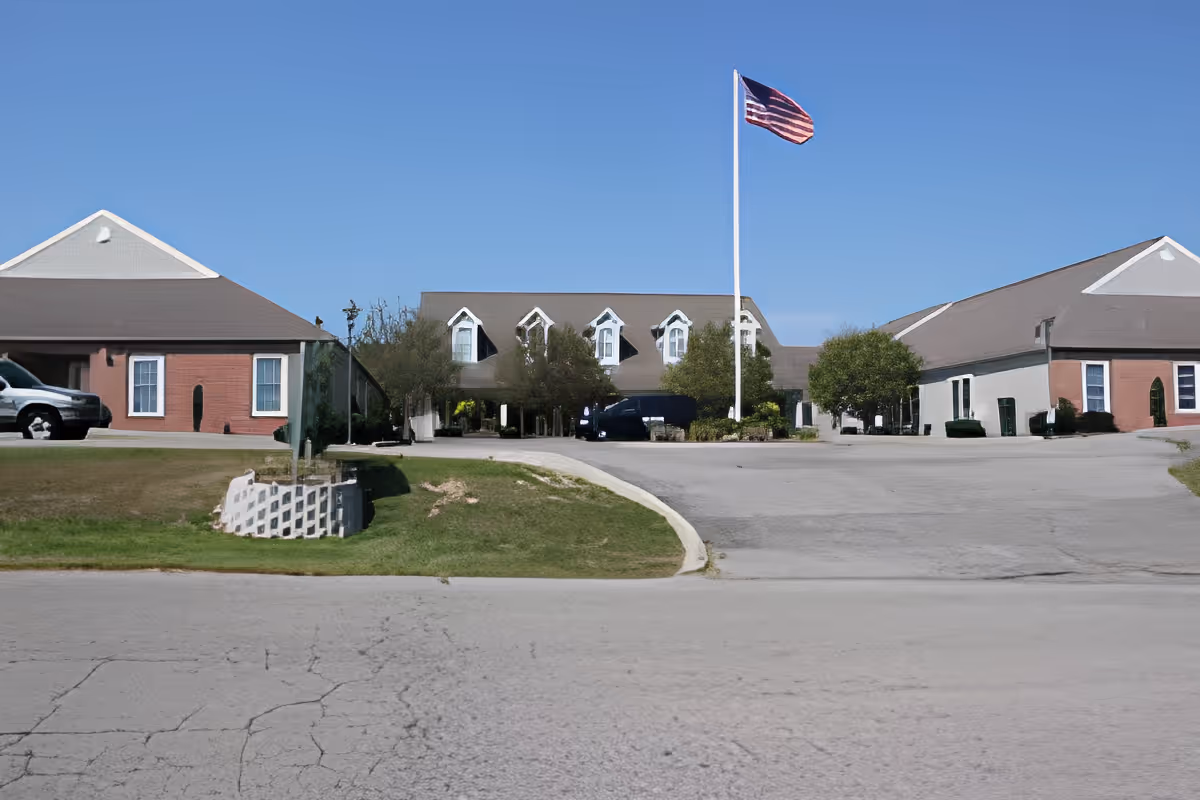 Exterior front view of Holzer Assisted Living - Jackson facility with a driveway, an American flag on a tall flagpole, and a clear blue sky. The building has a brown roof, brick and light-colored walls, and several windows. There are some trees and shrubs near the entrance.