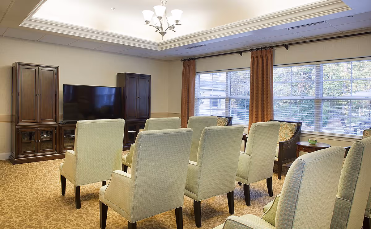 A cozy living room with six beige upholstered armchairs arranged in two rows facing a large flat-screen TV mounted between two wooden cabinets. The room has large windows with brown curtains allowing natural light to enter, and a chandelier hanging from the ceiling.