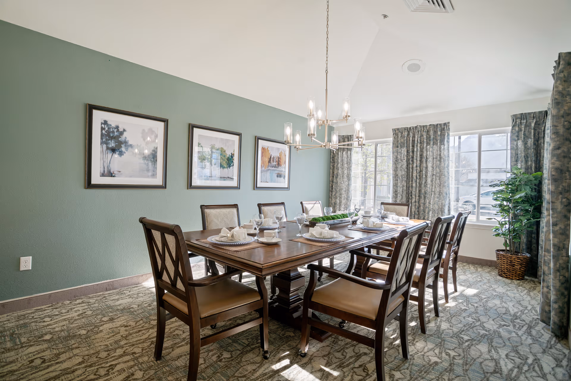 Sunlit dining room with a long wooden table set for eight, framed artwork on a green wall, and large windows with curtains.