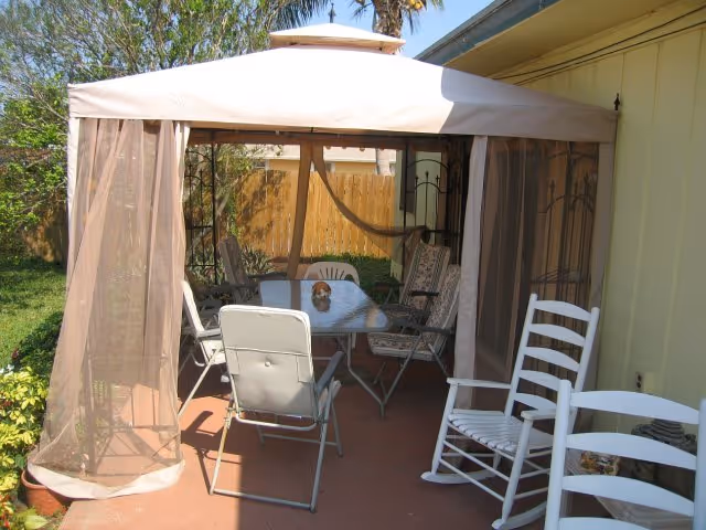 Outdoor patio area with a canopy tent featuring sheer curtains, a glass-top table surrounded by various chairs including a white rocking chair, and a wooden fence in the background with greenery around.